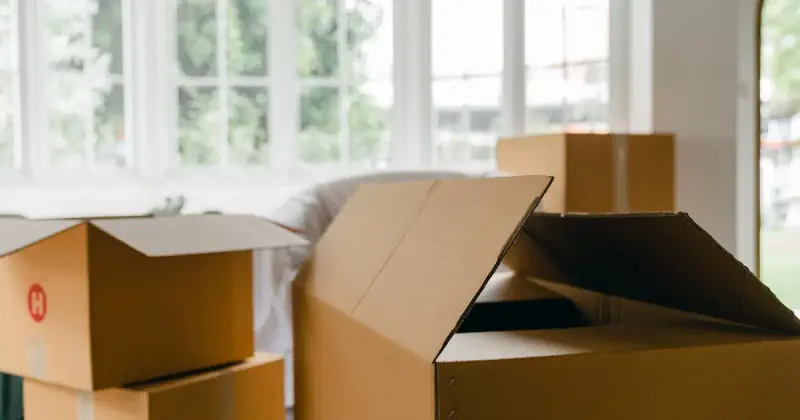 A woman carefully wraps a sofa in protective material while packing boxes in a bright room.