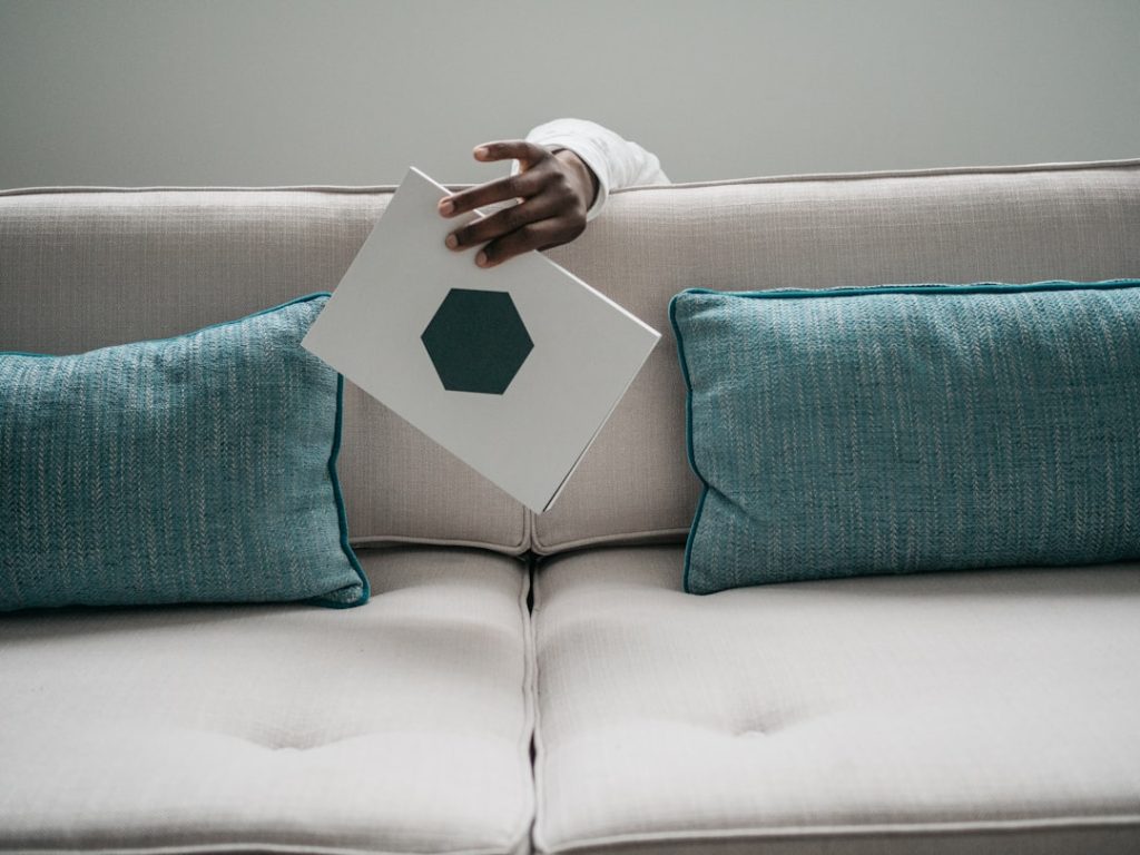 Hand holds white book on couch with pillows.