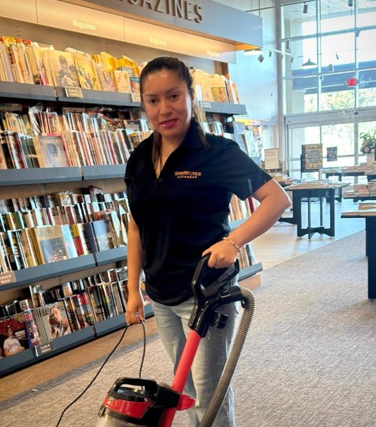 Residential photo 2 Smiling cleaning professional with vacuum, SparkTex Cleaners logo on shirt, in bookstore.